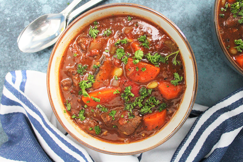 Thick, delicious, hot, Italian Beef Stew, filled with chunks of carrot, beef, beans, and tomato, topped with fresh herbs in a big bowl.