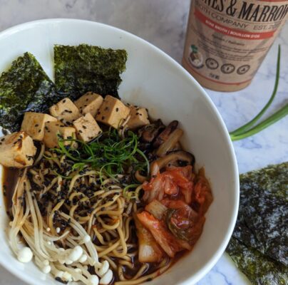 A big bowl of ramen topped with tofu, kimchi, mushrooms and seaweed, with Bones & Marrow Ramen broth in the background.